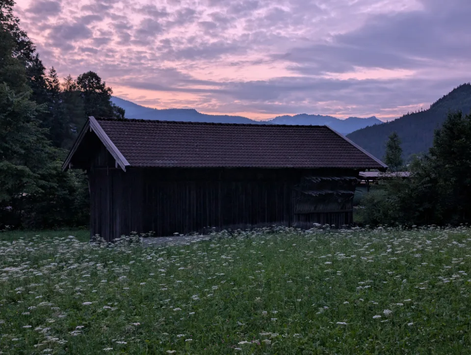 Alte Heuhütte auf einer Bergwiese in der Abenddämmerung, dahinter die Silhouette der Alpen